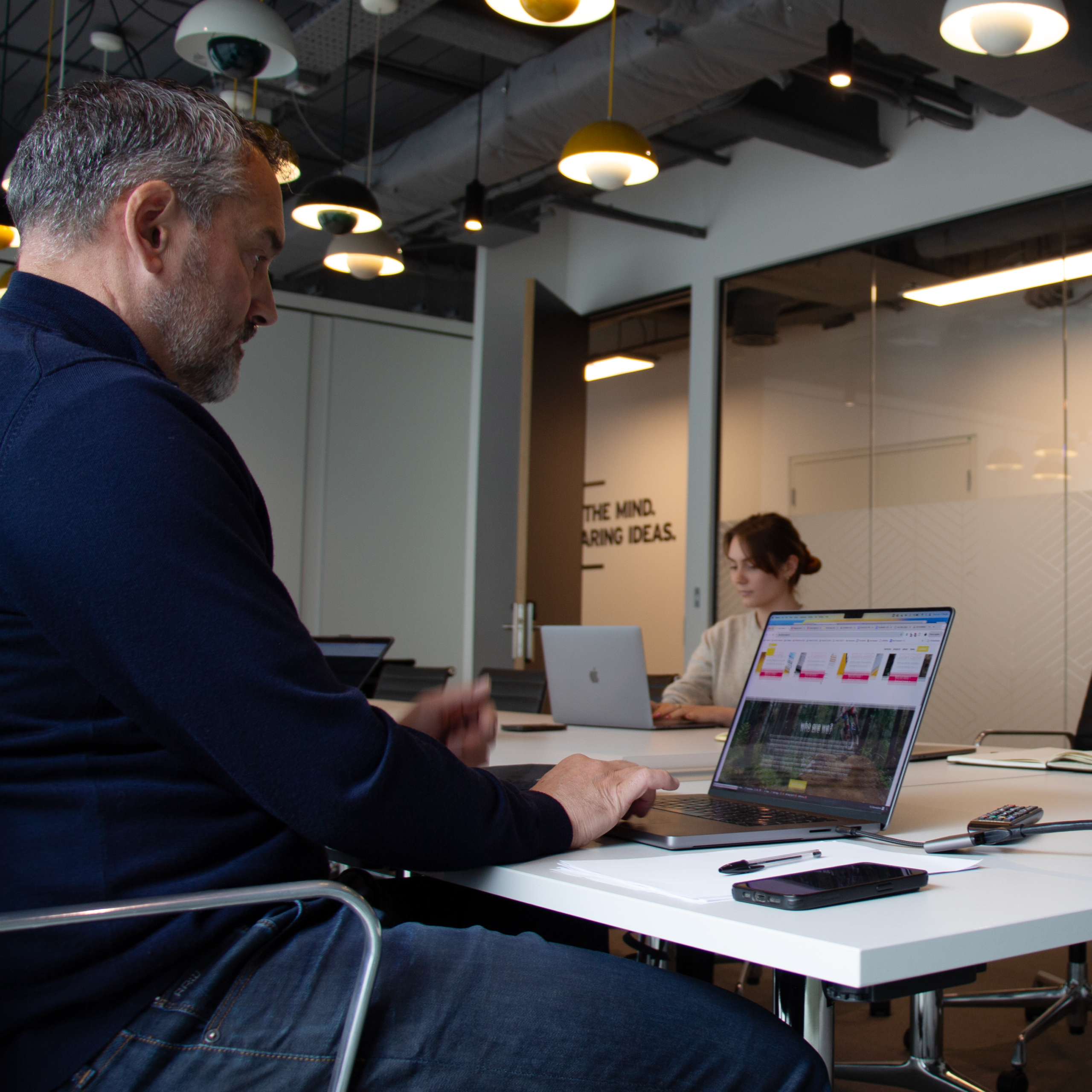 A man in a blue sweater working on a laptop at a long white conference table with a woman working in the background.