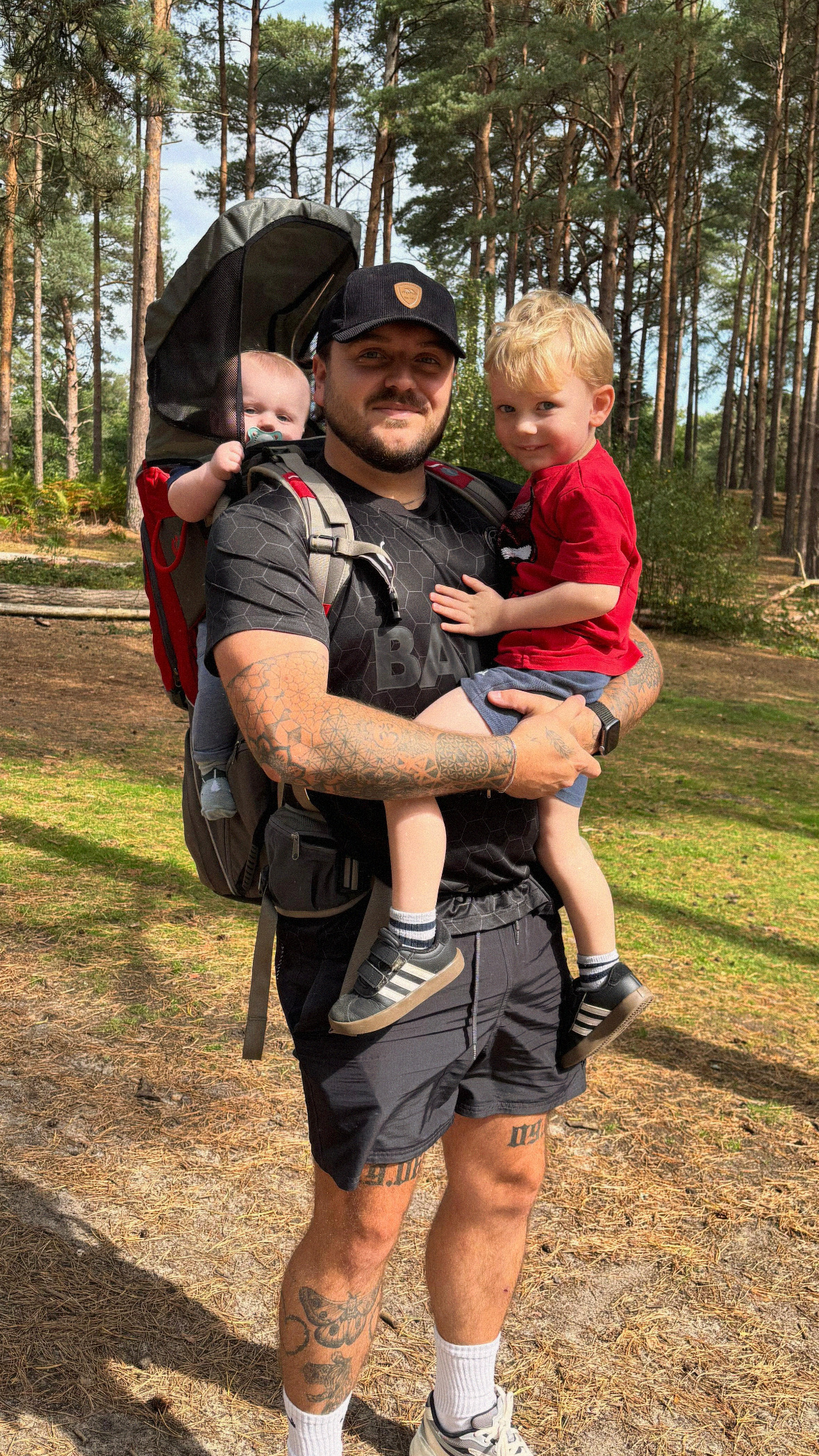 A man with tattoos on his arms holding a young child and carrying a baby in a hiking backpack in a wooded area.