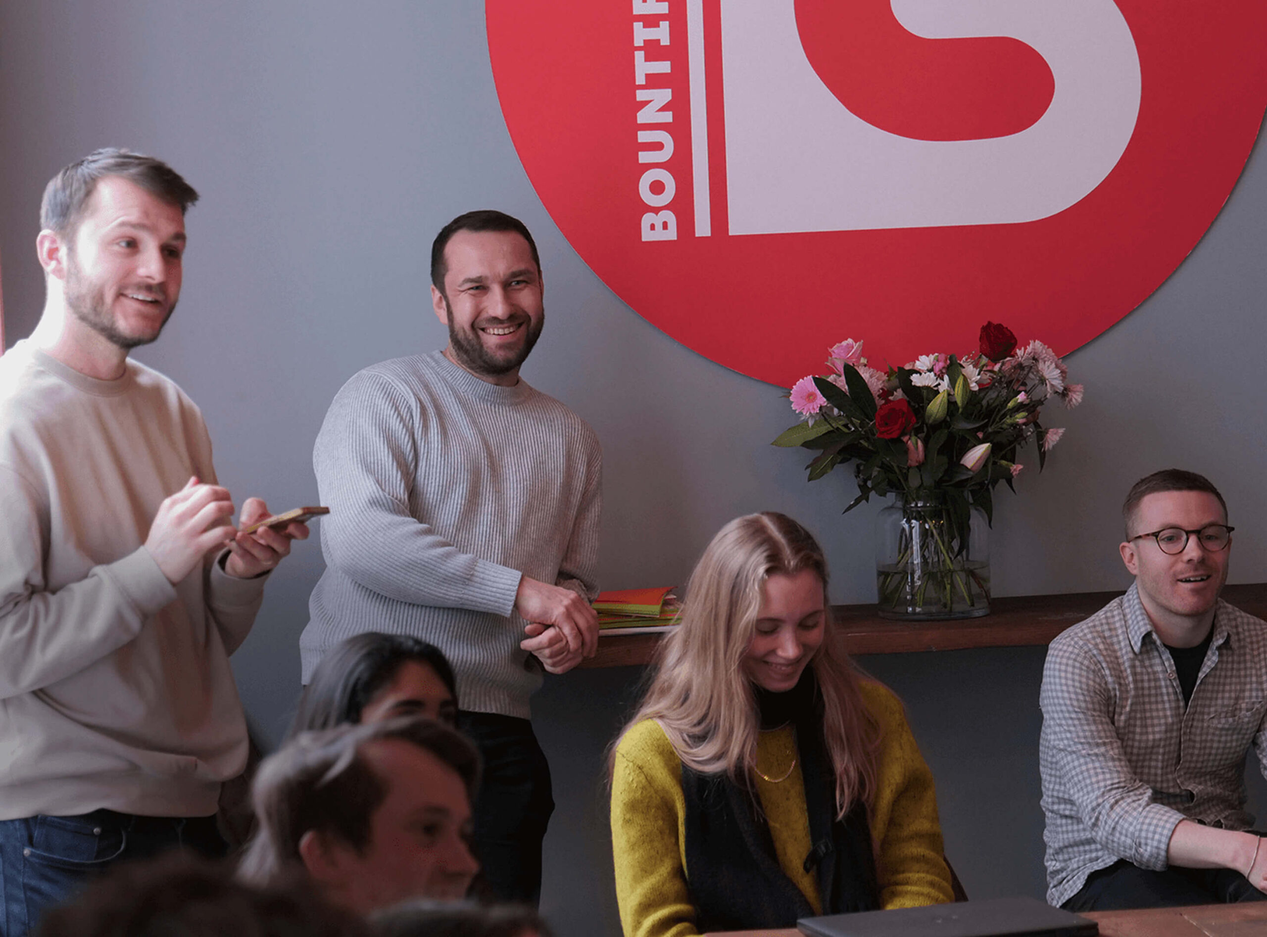 Group of smiling colleagues in a creative office space with a large red Bountiful Cow logo and flowers on a shelf