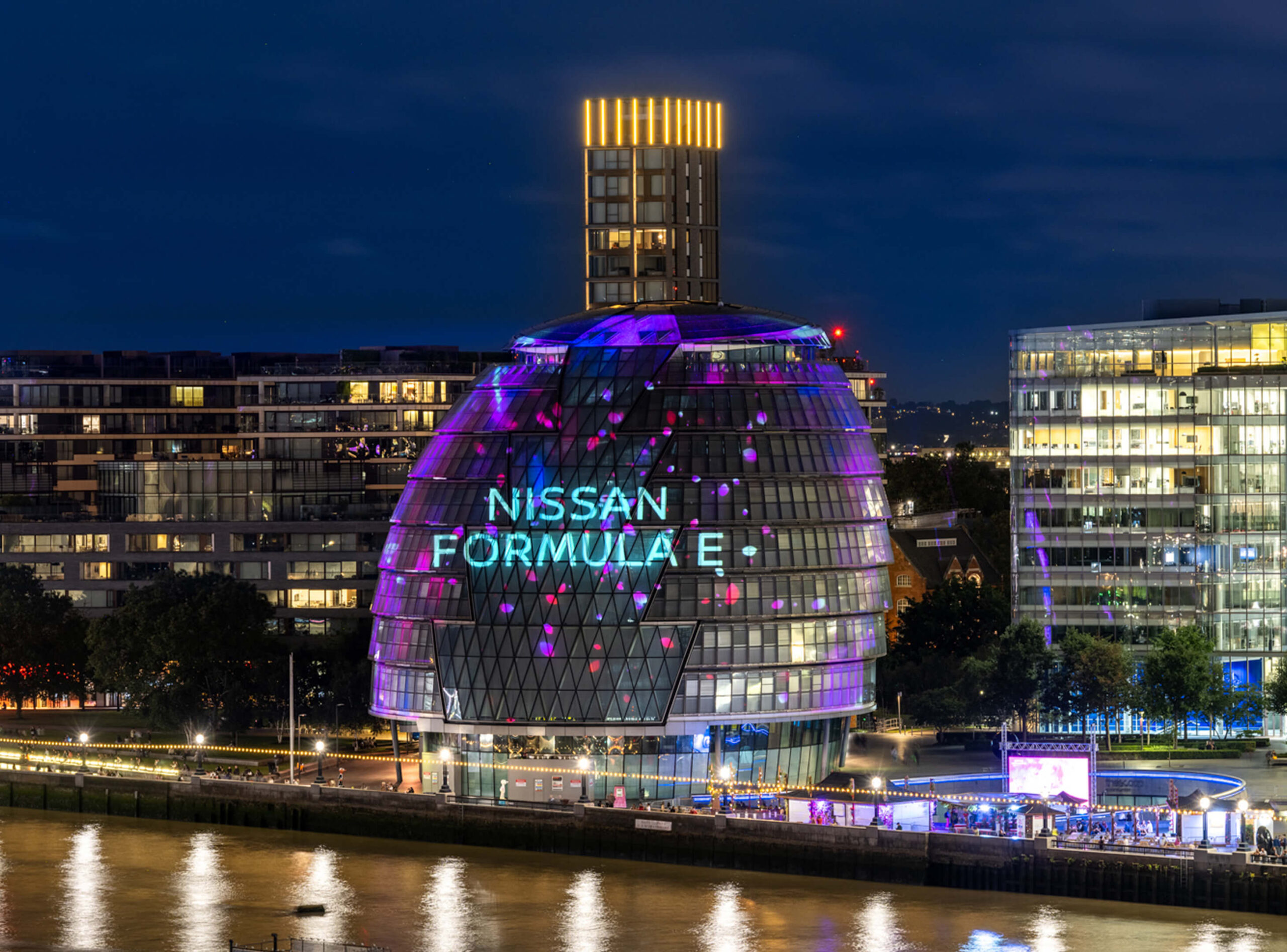 Nighttime cityscape with a modern glass building illuminated with Nissan Formula E lighting and neighbouring office buildings reflecting on the river.