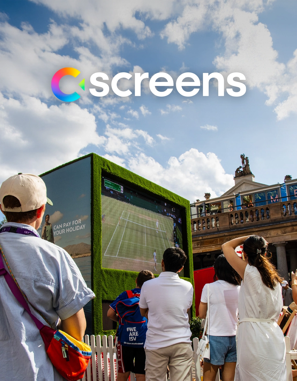 A group of people watching a tennis match on a large outdoor digital screen encased in artificial grass, with the C-screens logo overlaid on a bright blue sky.