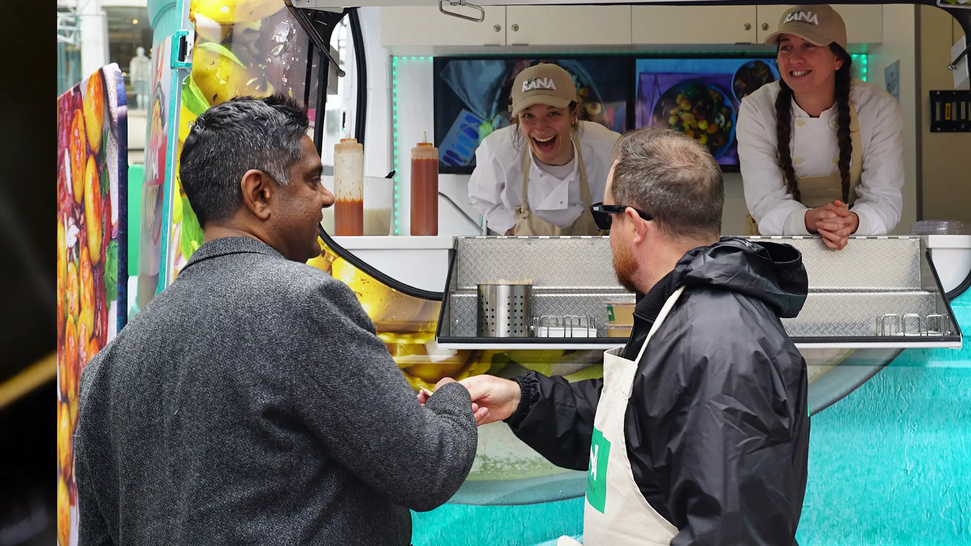 A man in a dark jacket stands in front of a colorful food truck, interacting with two smiling staff members wearing hats and white uniforms.