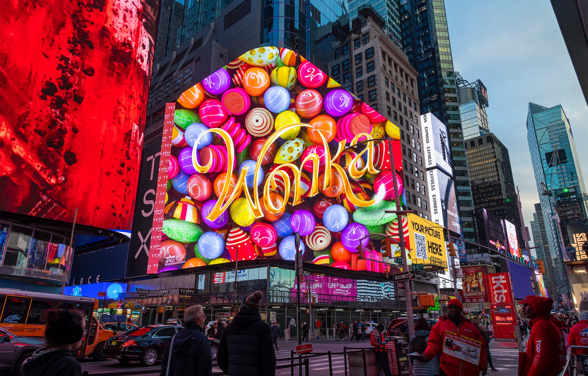 A massive digital billboard in a busy metropolitan square displays a vibrant, 3D-effect advertisement for "Wonka" featuring colourful sweets.
