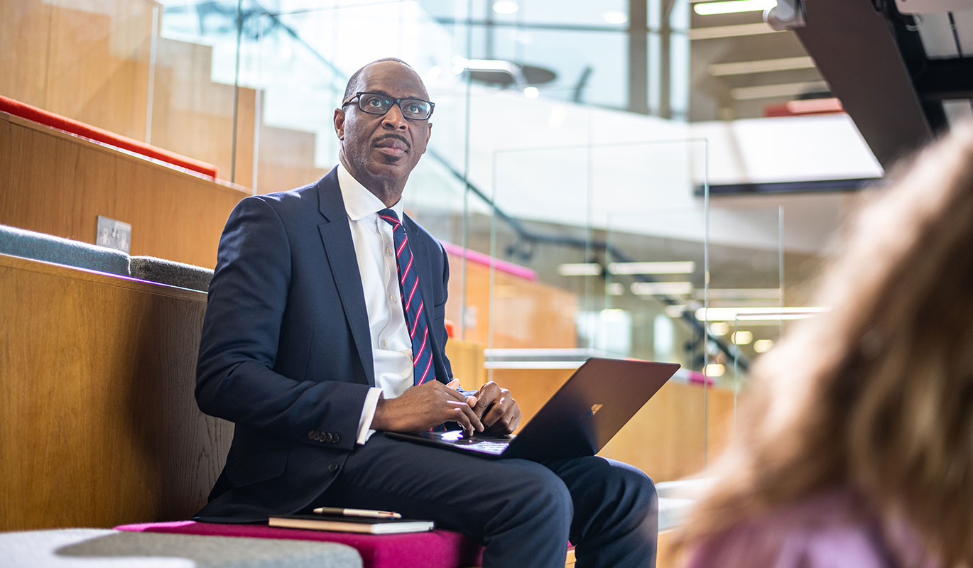 A man in a navy blue suit and glasses sitting in a modern, tiered seating area with a laptop open on his lap.