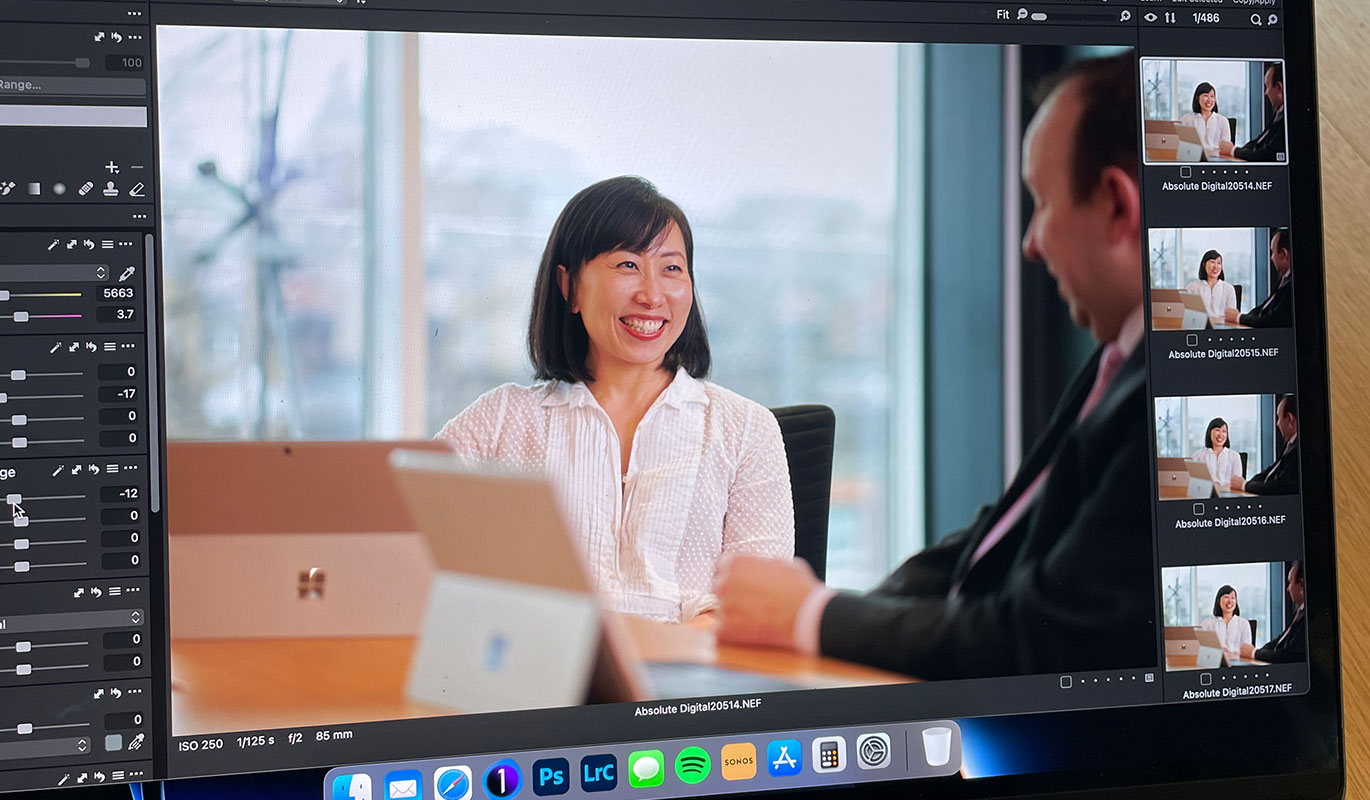 A laptop screen showing professional photo editing software with a smiling woman in an office setting being colour-graded.