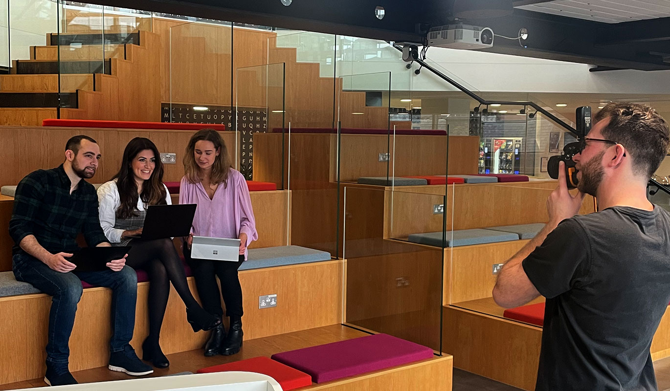 A videographer filming a group of three people sitting on a tiered wooden seating area in a modern office.