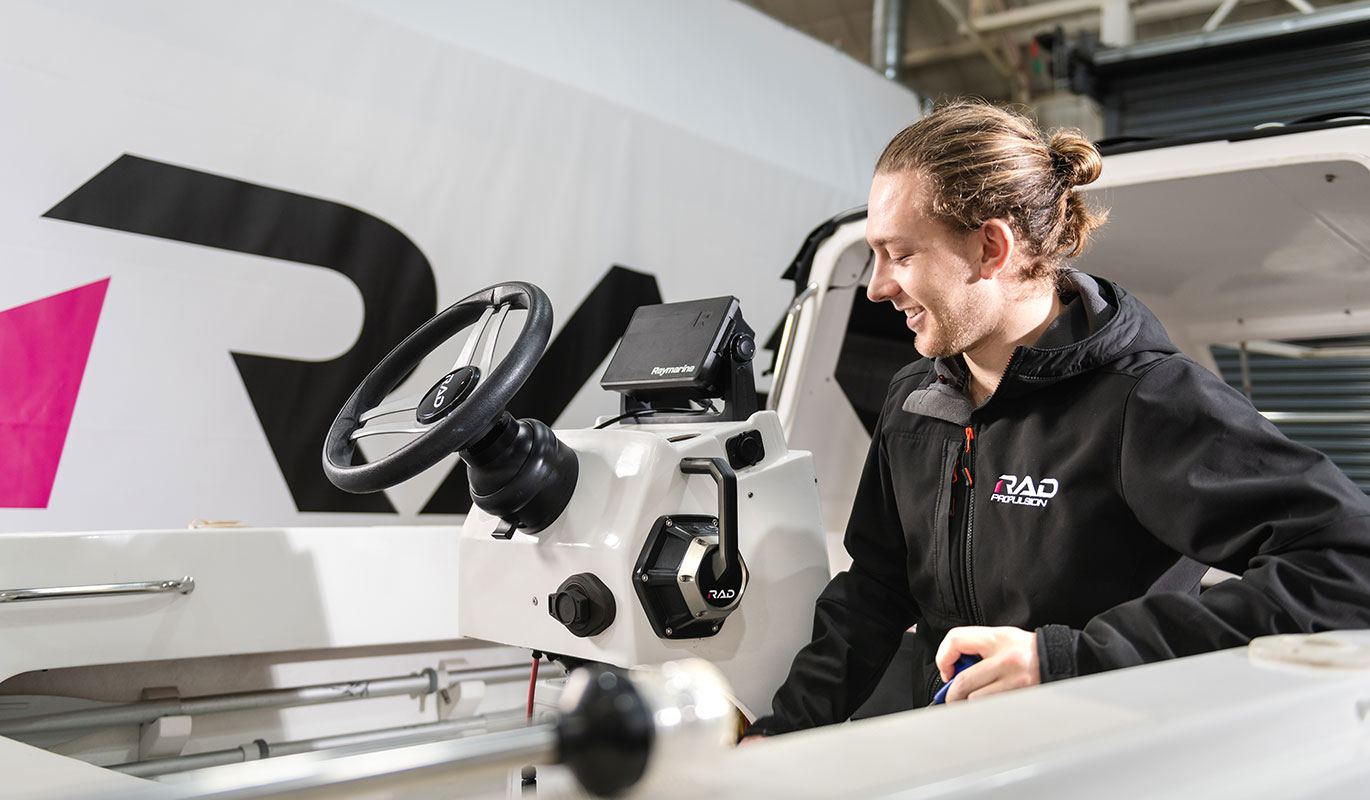 A man in a black jacket smiling while working on the steering console and navigation systems of a modern boat.