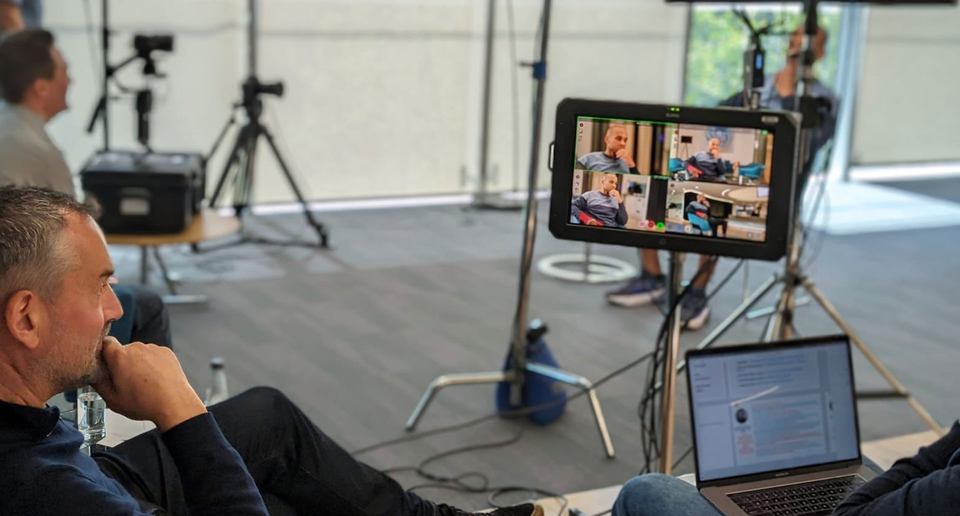 A behind-the-scenes view of a professional video shoot showing a production monitor, camera equipment on tripods, and a crew member reviewing a laptop.