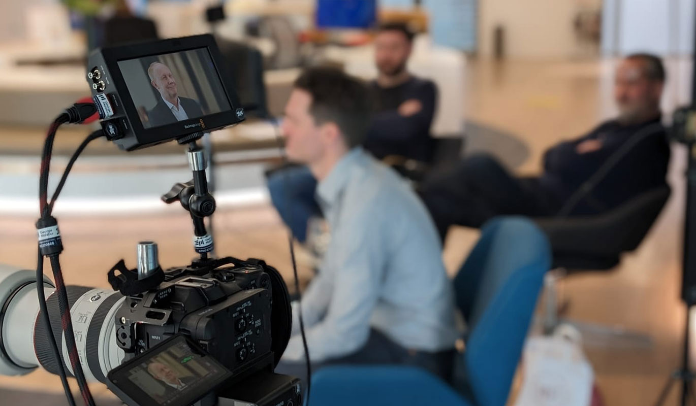 A close-up of a professional video camera monitor displaying a man being interviewed in a bright office setting