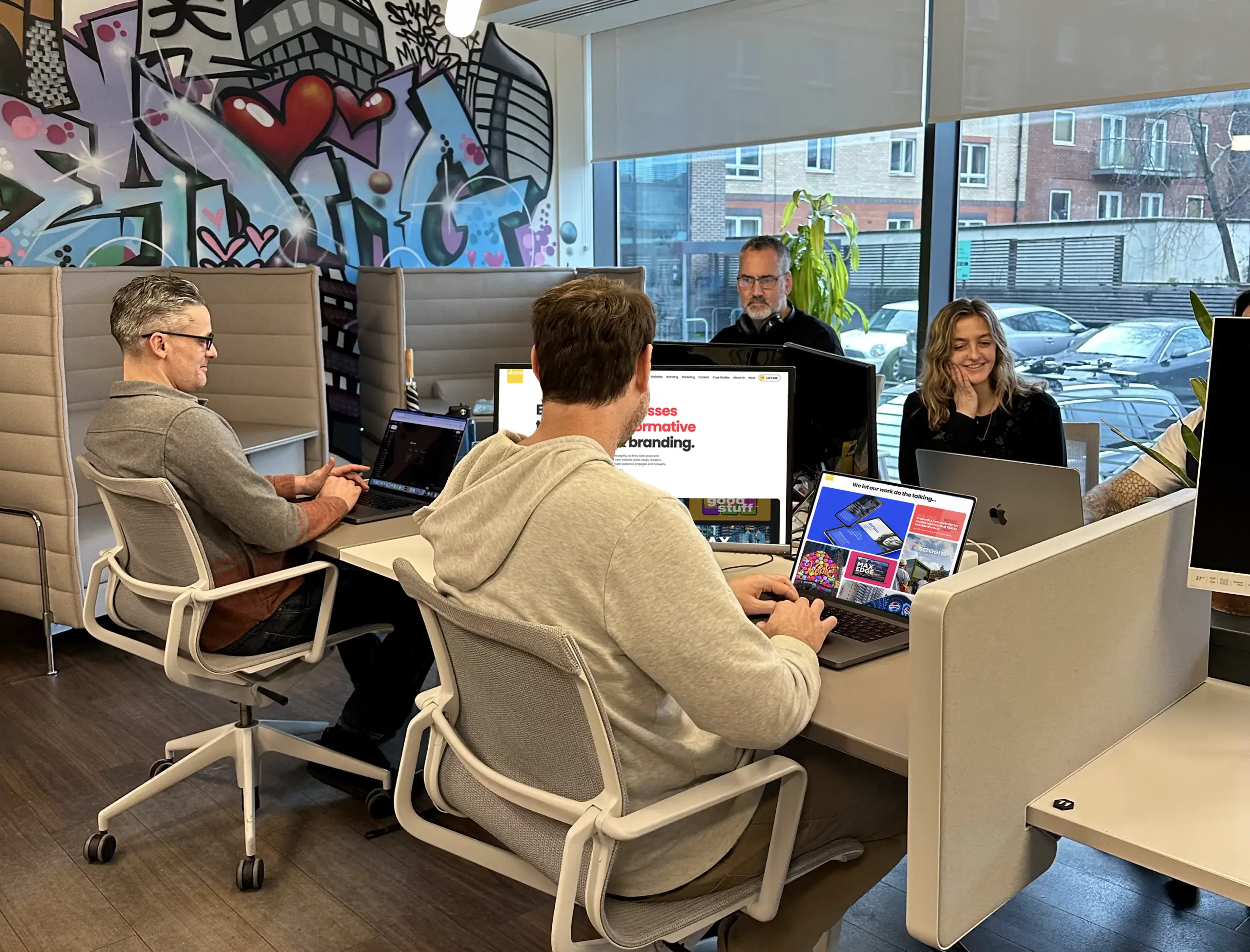 The Above Digital Team sit in-office, collaborating at desks, and smiling