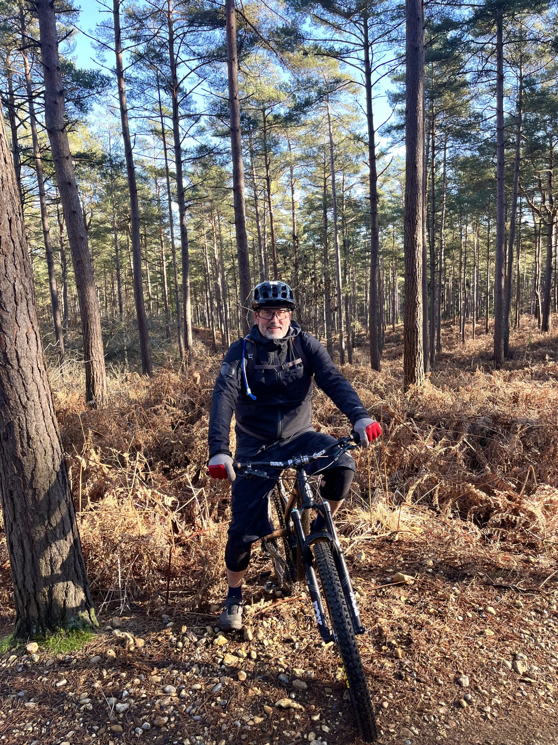 A man wearing a cycling helmet and gear posing with a mountain bike on a forest trail with tall pine trees.