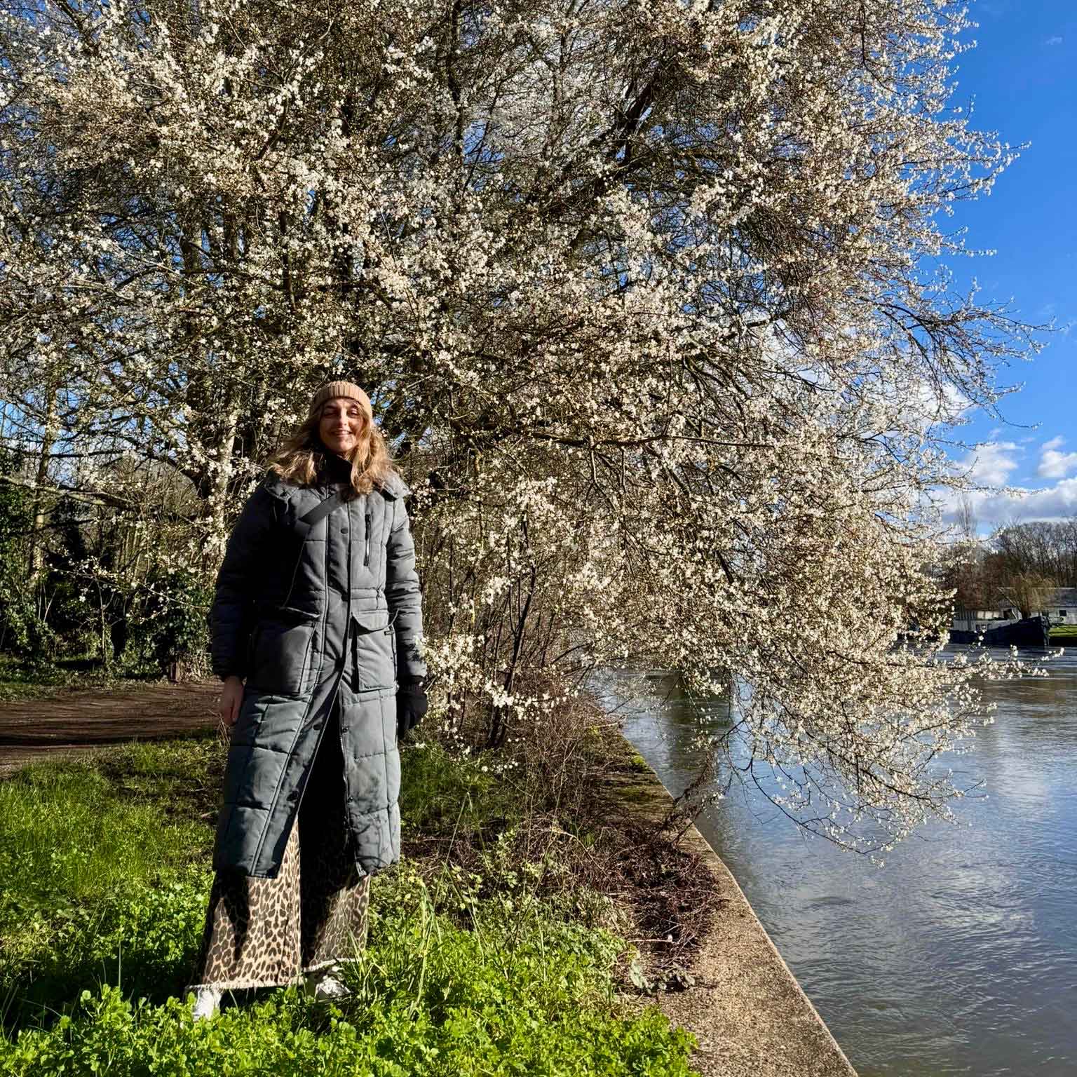 Smiling woman standing by a riverbank beside a tree in blossom on a cold sunny day.